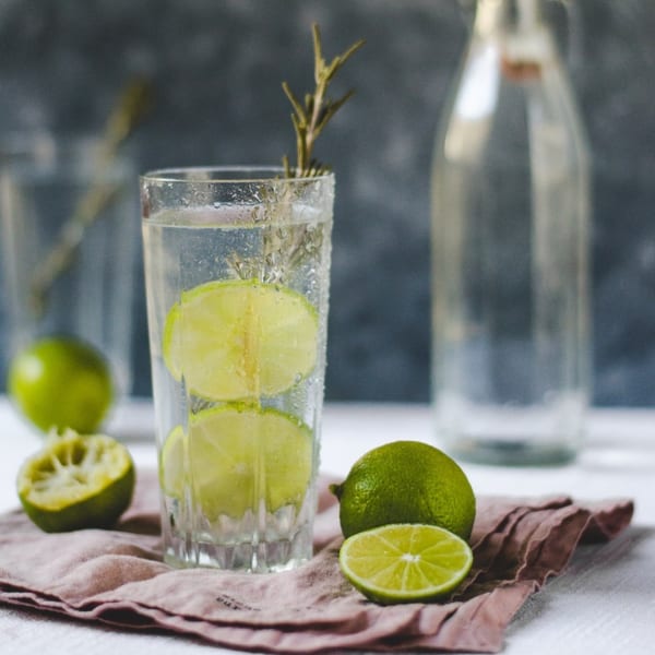 Refreshing lime and rosemary drink in a tall glass, with whole and sliced limes on a pink napkin.