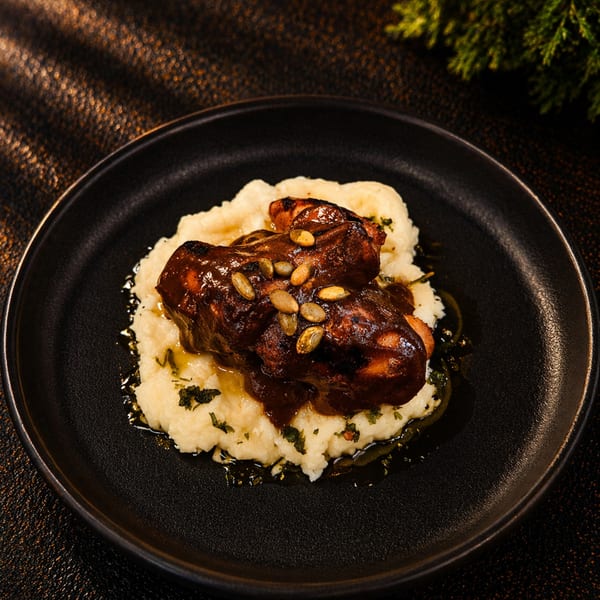 Braised short ribs with dark sauce and pumpkin seeds on a bed of mashed potatoes, garnished with herbs.