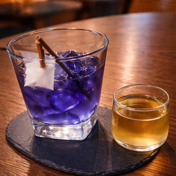 A vibrant purple cocktail with a paper boat garnish next to a small glass of amber liquid.