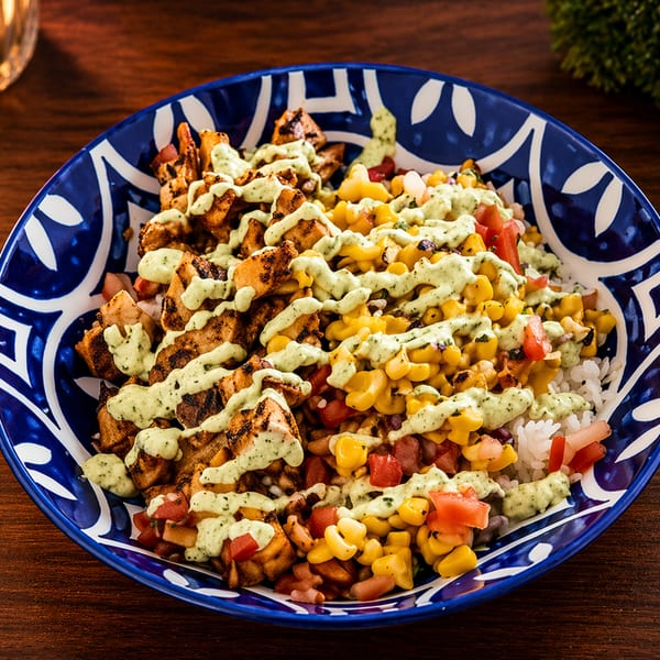 Grilled chicken, corn, and diced tomatoes over rice, drizzled with green sauce in a blue and white bowl.