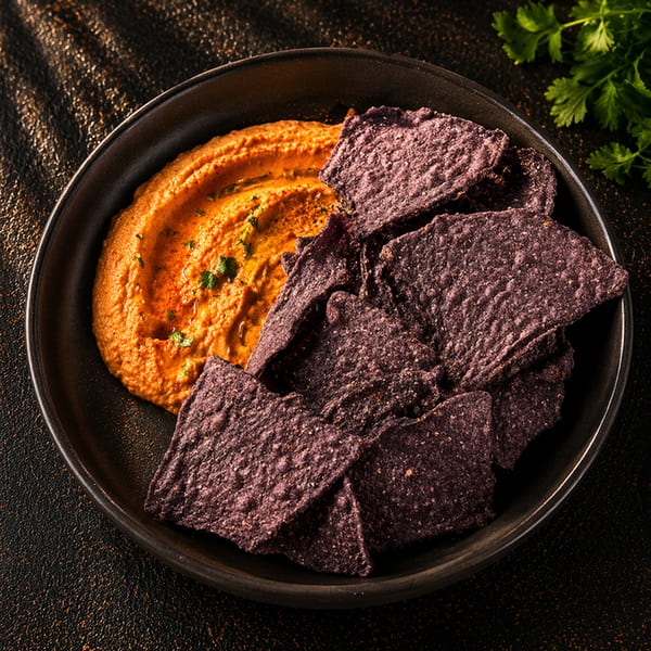A black bowl filled with vibrant orange hummus and a pile of dark purple tortilla chips.